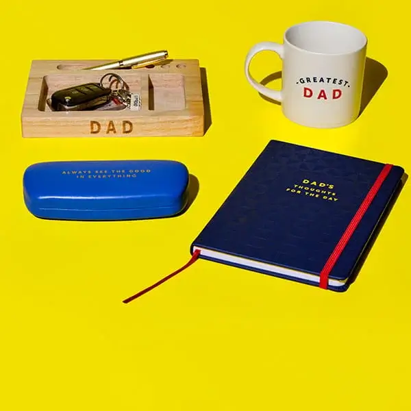 Fathers Day gifts on a yellow background including a personalised wooden desk tray, greatest dad mug, notebook, glasses case, and keys